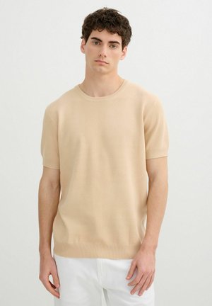 Joven con cabello corto, rizado y oscuro, que lleva un suéter de punto beige de manga corta y pantalones blancos sobre un fondo claro y liso.