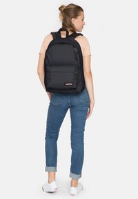Sac à dos en tissu noir avec des bretelles rembourrées, poche zippée à l'avant et patch de logo. L'utilisateur porte une chemise claire et un jean. Design simple et fonctionnel.