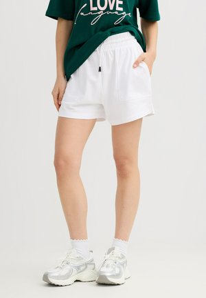 Mujer que lleva shorts blancos con cintura elástica, camiseta verde con el texto "LOVE language", calcetines blancos hasta el tobillo y zapatillas plateado-blancas, de pie con una mano en el bolsillo.