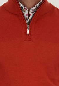 Rode zip-up trui met een structuurbreipatroon, voorzien van een hoge kraag en een metalen rits. Daaronder is een bloempatronen shirt zichtbaar.