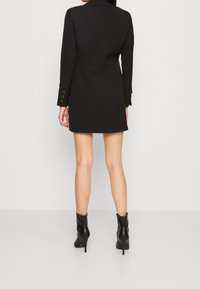 Vestido mini negro entallado tipo blazer con un diseño estructurado, mangas largas y acentos de botones dorados en los puños. Combinado con botas negras hasta el tobillo.