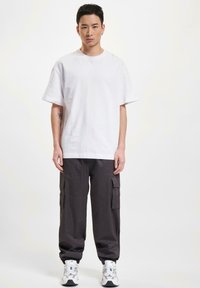 Hvid oversized t-shirt parret med mørkegrå cargo bukser, der har sidelommer, fuldendt med hvide og teal sneakers. Ensfarvet baggrund.