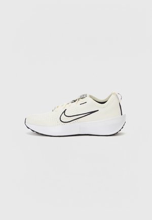 Light beige athletic shoe with a textured upper, black swoosh logo, and a white cushioned sole. Features lace-up design and padded collar.
