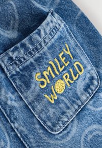 Sininen farkkukangas, jossa on ommeltu tasku, jossa on keltainen brodeeraus, jossa lukee "SMILEY WORLD". Kangas on pehmeä ja siinä on haalistunut kuvio.