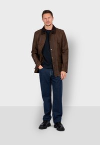 Chaqueta de cuero marrón con un cuello más oscuro, cierre de cremallera y botones a presión; llevada sobre un suéter negro y jeans azules, combinada con zapatos negros.