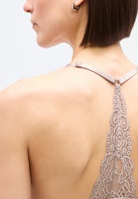 Κοντινό πλάνο της πλάτης μιας γυναίκας με κοντά καστανά μαλλιά, που φοράει ένα φόρεμα με τιράντες, με δαντελένια κάθετη τιράντα και ένα μικρό μαύρο σκουλαρίκι.