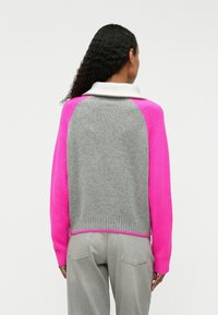 Pull en tricot avec un corps gris et des manches roses, doté d'un col blanc côtelé. Texture tricotée avec des couleurs contrastées et des détails de motif.