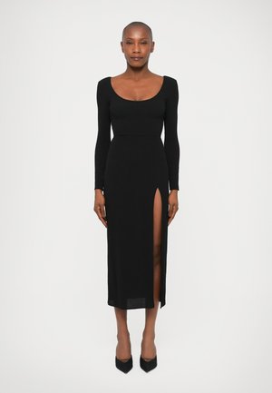 Lungo vestito nero in un design aderente, con scollatura a barca e uno spacco alto sulla coscia; realizzato in un materiale liscio e elasticizzato.