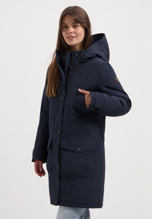 Vrouw in een marineblauwe winterjas met capuchon, grote voorzakken, knopen- en ritssluiting, staande tegen een effen achtergrond.