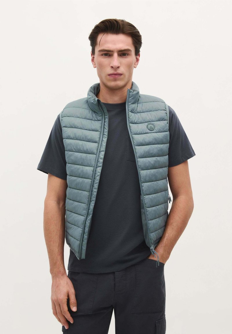 Gevoerde vest in gedempt groen, met een hoge kraag, ritssluiting en horizontale stiksels. Draag het over een donker T-shirt met korte mouwen.