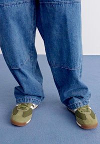 Tênis verdes e cinzas com detalhes brancos, combinados com calças de denim azuis largas, apresentando uma textura desbotada e costuras visíveis.
