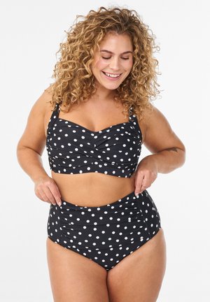 Smiling woman with curly hair adjusts black and white polka dot high-waisted bikini against plain white background.