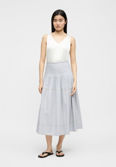 Femme debout, portant un haut blanc sans manches, une jupe midi à rayures bleu clair taille haute, et des tongs noires, sur un fond uni.