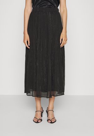 Femme portant une jupe midi plissée noire avec des rayures verticales argentées et des sandales à talons ouvertes noires, debout devant un fond blanc.