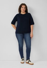 Marineblauer kurzärmeliger Strickpullover mit rundem Ausschnitt, kombiniert mit dunkelblauen Jeans und weißen Turnschuhen mit schwarzen und braunen Akzenten.