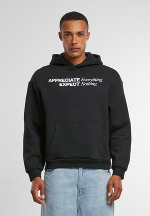 Zwarte hoodie van zacht materiaal, met een kangoeroe-zak, verstelbare capuchon en grote tekst aan de voorkant: "APPRECIATE EXPECT Nothing."