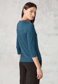 Türkisfarbener gestrickter Oberteil mit geometrischem Muster, 3/4-Ärmeln und rundem Kragen, kombiniert mit schwarzen Hosen.