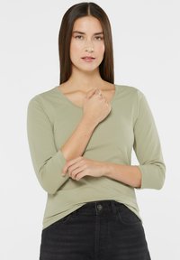 Khaki green long-sleeve top with a rounded neckline and fitted shape. Soft fabric and three-quarter sleeves. Paired with black pants.