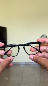 Armação de óculos em plástico preto com formato de olho de gato. Apresenta detalhes em prata nos dobradiças e uma gravação que diz "PARA DIAS NUBLADOS E HUN."