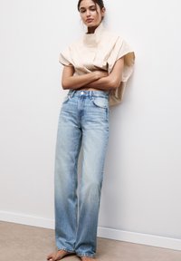 Svetlo bež oversized majica z visokim ovratnikom, v kombinaciji s srednje visokimi širokimi faded modrimi kavbojkami. Gladek material in denimska tekstura.