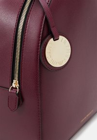 Vinröd texturerad läderväska med en guldfärgad dragkedja, cirkulär bricka och präglad logotypdetalj på en slät yta.
