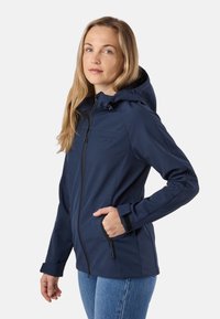 Femme aux longs cheveux blonds portant une veste à capuche bleu marine zippée et un jean bleu, avec une main dans la poche.