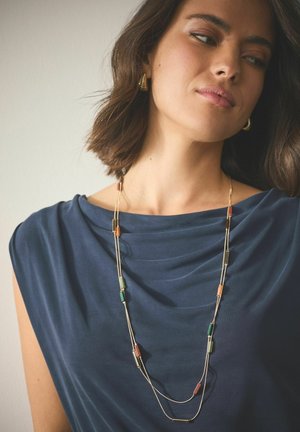 Goldfarbene Lagenkette mit rechteckigen Perlen in Orange, Grün und Rot, getragen über einem marineblauen, drapierten Oberteil.