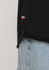 Schwarzer Hemdsaum mit kleinem rot, weiß und blauem Etikett über der Naht der Tasche einer hellblauen Jeans.