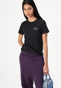 T-shirt de algodão preta com um pequeno logótipo rosa "Converse" no peito. Usada com calças de treino roxas e uma bolsa azul.