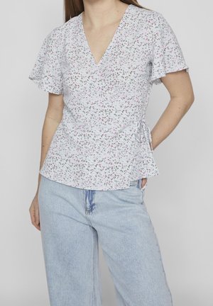 Vrouw draagt een witte, bloemige korte-mouwen wikkelblouse met V-hals en lichtblauwe hooggetailleerde jeans tegen een effen achtergrond.