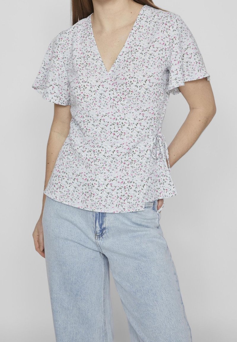 Femme portant un chemisier cache-cœur blanc à manches courtes fleuri avec un col en V et un jean taille haute bleu clair sur un fond uni.