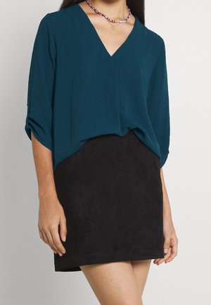 Blouse en viscose teal avec un col en V et des manches roulées, associée à une mini-jupe en daim noir. Le tissu est doux avec un léger éclat.