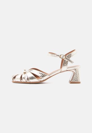 Metāliska sudraba sandales ar augstu papēdi, teksturētu virsmu, atvērtu purngalu, krusti savilktām siksnām, regulējamu potītes siksnu un biezām papēžiem.