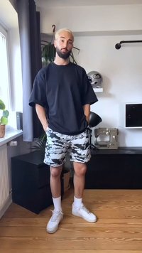Man with blonde hair and beard stands indoors wearing black oversized t-shirt, black and white camouflage shorts, white socks, and white sneakers.