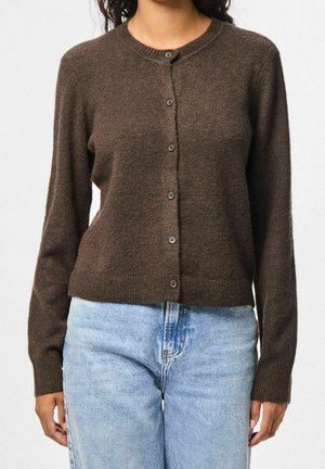Vrouw draagt een aansluitend bruin vest met knopen en lange mouwen, gecombineerd met lichtblauwe hoog in de taille vallende jeans.