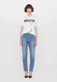 Balta medvilninė marškinėlių su piešiniu, kurioje pavaizduotas antis ir užrašas "MOSCHINO", derinami su šviesiai mėlynomis džinsinėmis kelnėmis ir juodais smailiais aukštakulniais.