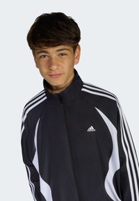 Dospievajúci chlapec s tmavými vlasmi, oblečený v čierno-bielej športovej bunde Adidas so zipsom, pozerajúci sa mierne do strán s neutrálne výrazom tváre.