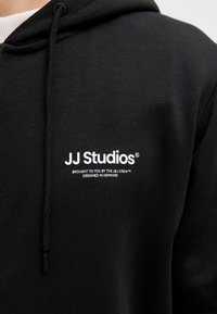 Schwarzer Hoodie mit strukturiertem Finish. Verfügt über eine Vordertasche, eine Kapuze mit Kordelzug und weiße Logos: "JJ Studios" sowie zusätzlichen Text darunter.