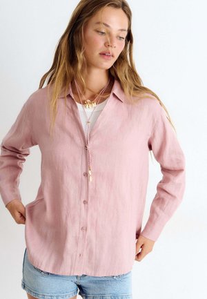 Femme aux longs cheveux porte une chemise rose ample boutonnée sur un haut blanc, un short en jean et des colliers superposés avec des détails coquillages.