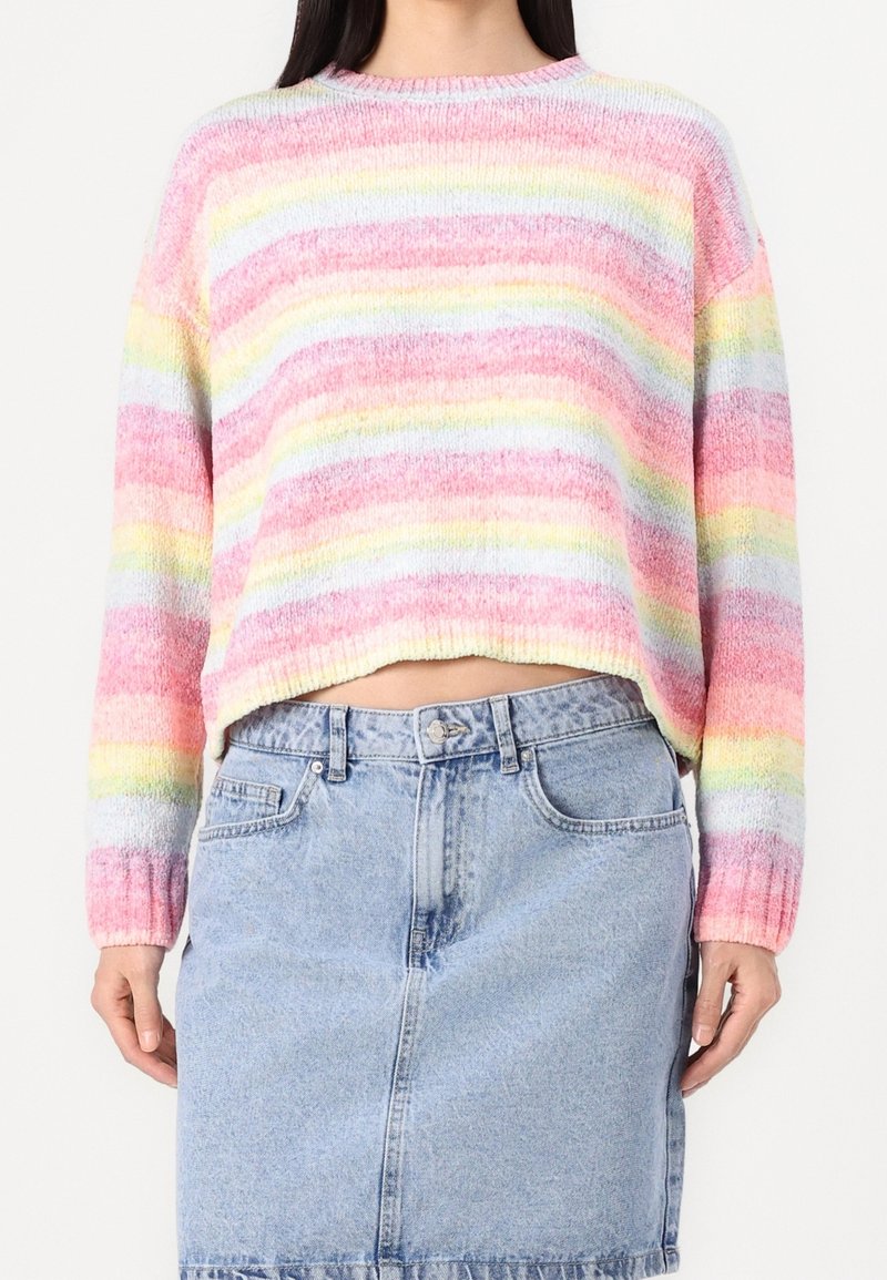 Femme portant un pull doux rayé multicolore pastel et une jupe en denim bleu clair, debout devant un fond blanc uni.