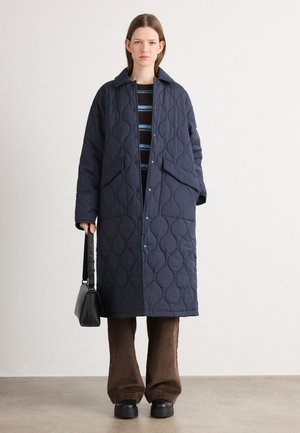 Marineblauwe, gewatteerde jas met een kraag, voorstaande drukknoopsluiting, zijzakken en een relaxte pasvorm, gedragen over een gestreepte trui en bruine broek.