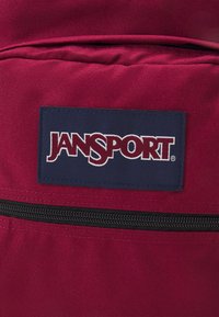 Crvena tkanina ruksak s pravokutnom Jansport logotipom u tamnoplavoj boji i crnim zatvaračem. Tekstura izgleda glatko s ojačanim šavovima.