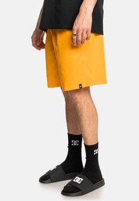 Helle orange Shorts aus glattem Stoff, knielang, mit einem kleinen schwarzen Logoschild. Kombiniert mit schwarzen Rutschsandalen und Socken.