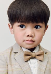 Enfant en costume gris clair à carreaux avec un nœud papillon beige, chemise blanche. Caractéristiques : texture lisse, coupe structurée et encolure arrondie. Yeux bleu vif.