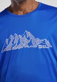 T-shirt de sport bleu avec un dessin linéaire blanc de montagnes et un logo en bas à droite. Texture lisse et design à col rond.