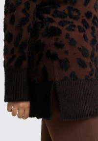Brauner Pullover mit schwarzen Leopardenflecken, strukturierter Stoff, gerippte Bündchen und Seitenöffnungen, getragen über braunen Hosen. Weiches, pelziges Finish.