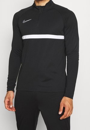 Homme portant un haut de sport noir à manches longues Nike avec une bande horizontale blanche sur la poitrine et une fermeture éclair quart de tour au col.