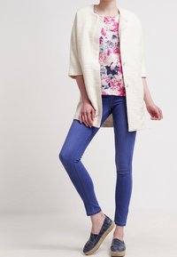 Veste texturée beige, top à imprimé floral, jean skinny bleu et chaussures navy à enfiler. Caractéristiques : manches courtes et fermeture à boutons.