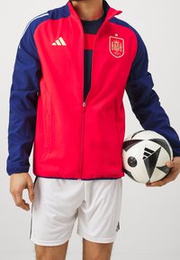 Red and blue, zippered jacket with the Spain national emblem. Worn with white shorts, holding a black and white soccer ball.