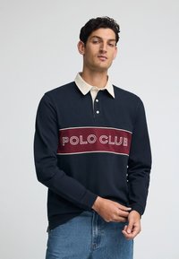 Granatowa koszulka polo z długim rękawem z kremowym kołnierzykiem, z wyrazistym bordowym logiem "POLO CLUB" na piersi, wykonana z bawełnianej tkaniny.